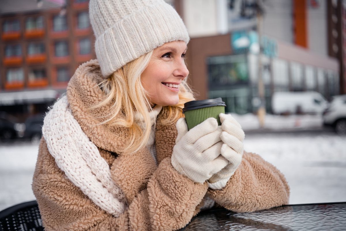 Riscaldarsi in inverno: donna beve un caffè caldo, indossando cappotto, sciarpa e guanti.