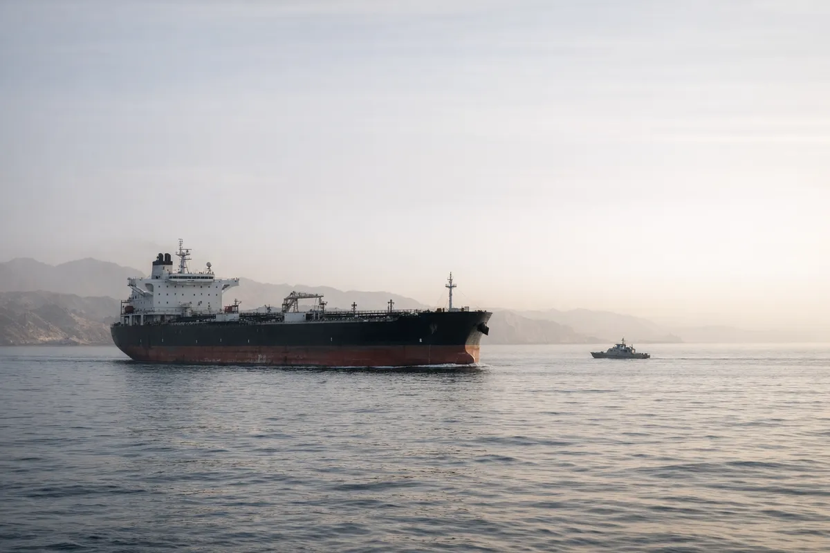 Petroliera in navigazione su mare calmo con piccola motovedetta sullo sfondo e costa montuosa nella foschia